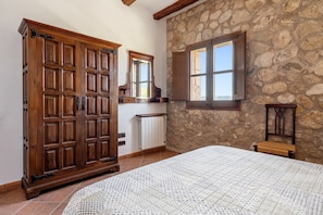 A bedroom with a large wooden wardrobe, a stone wall, a window, and a bed with a patterned bedspread.