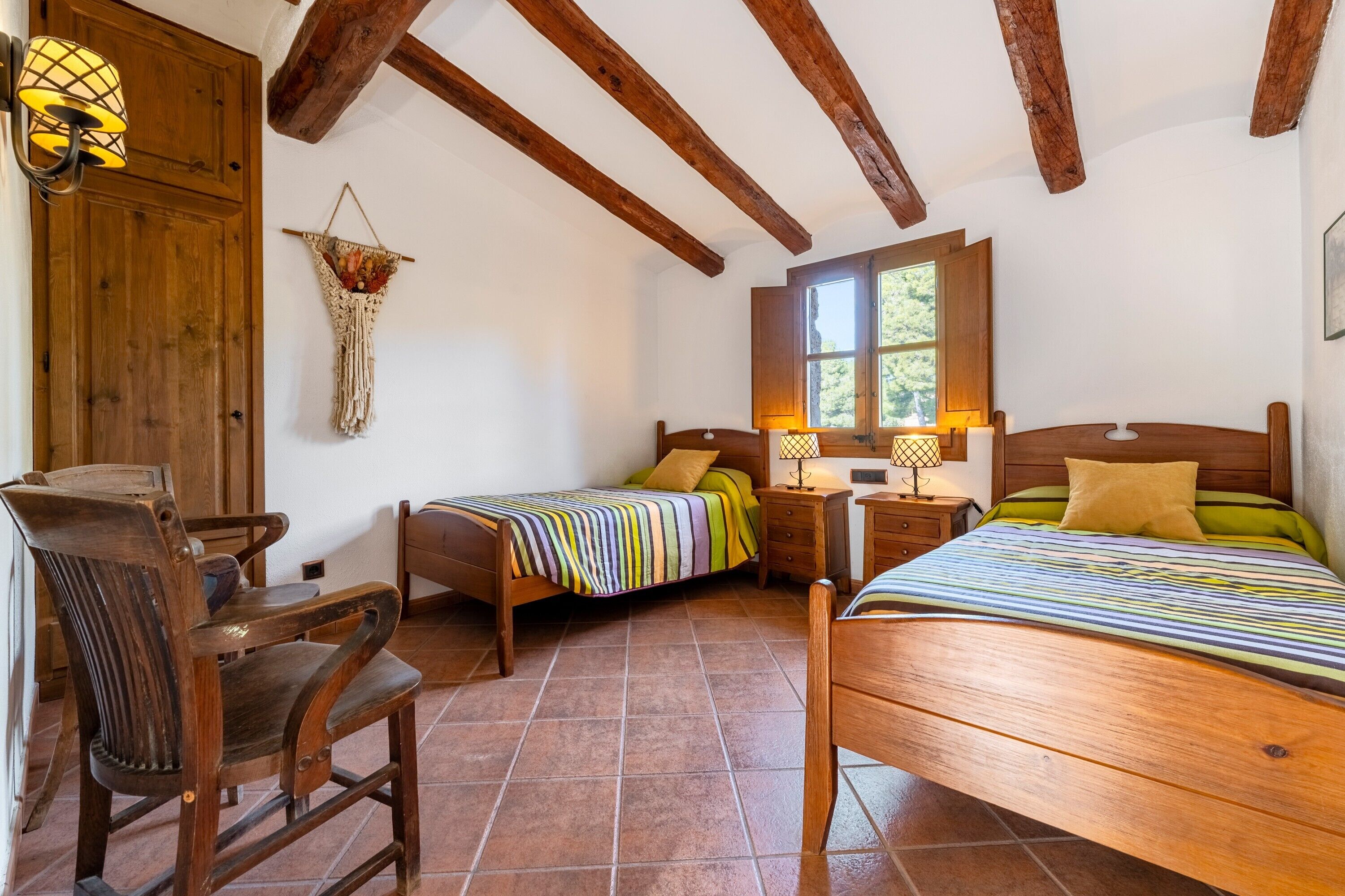 Una habitación con dos camas de madera, una silla de madera, un escritorio de madera, un adorno macramé en la pared y una ventana con luz natural.