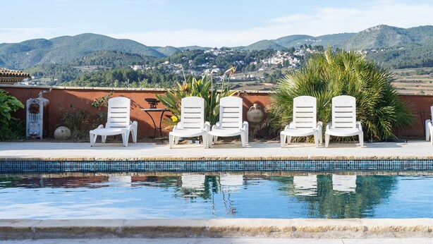 Una piscina con sillas reclinables blancas, borde de baldosas y vistas a montañas y casas al fondo.