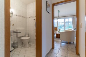 Een badkamer met toilet, wastafel en douche. Een gang leidt naar een woonruimte met een stoel en een tafel.