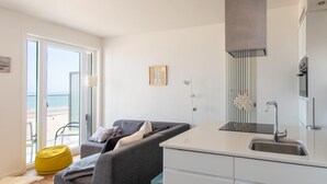 Une cuisine moderne avec un canapé gris et un fauteuil en forme de poire jaune, une grande fenêtre donnant sur la plage et un évier en acier inoxydable.