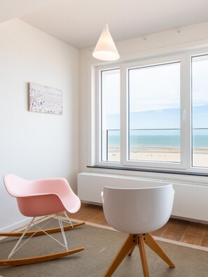 Una habitación con un amplio ventanal con vistas a la playa, una mecedora rosa, una silla blanca y una lámpara colgante blanca.
