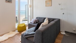 Un salon moderne avec un canapé gris, un fauteuil en forme de poire jaune et une vue sur la plage, visible à travers une porte vitrée.