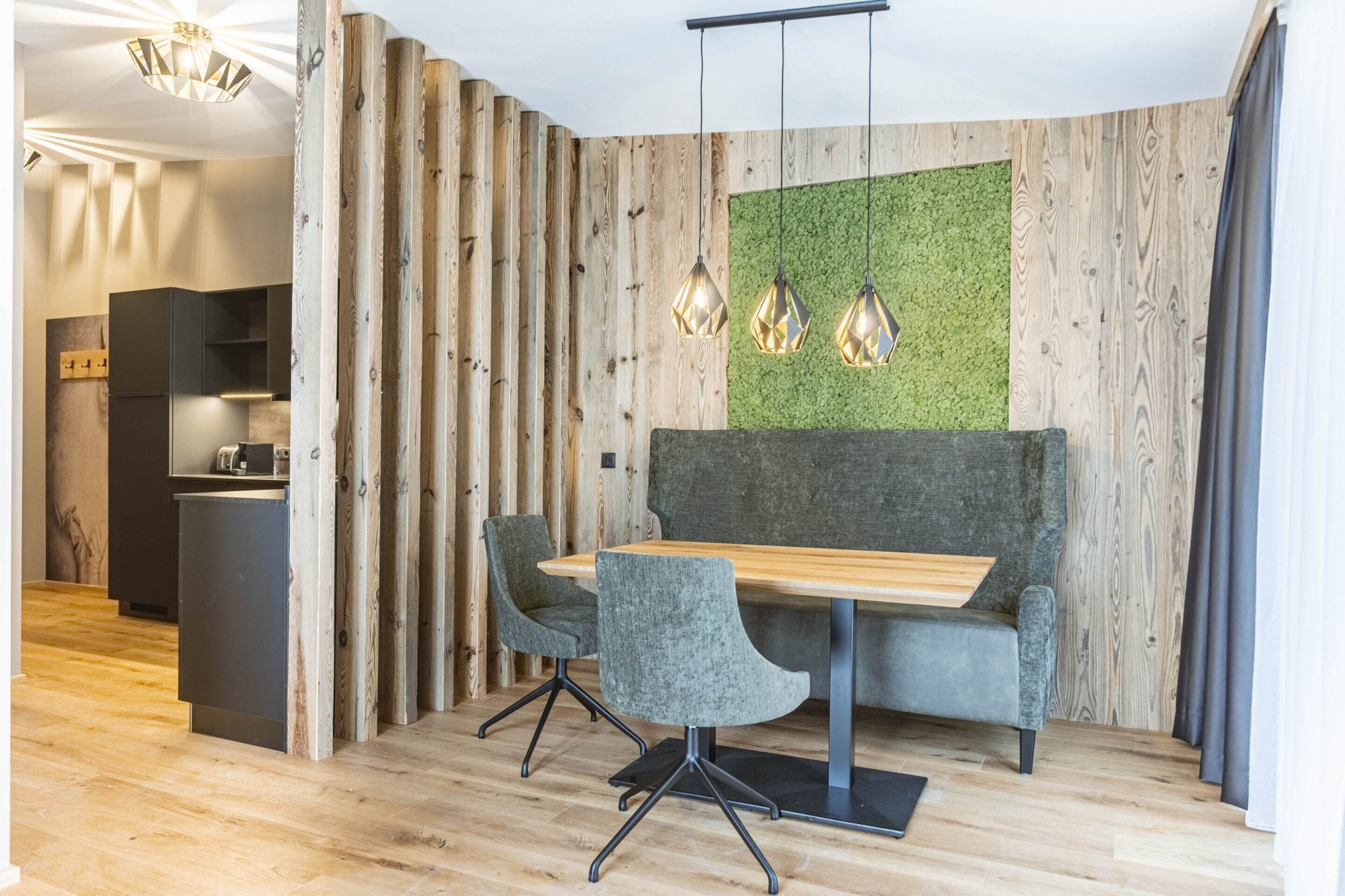 A modern dining area with a wooden table and two grey upholstered chairs, a green wall, and a kitchen in the background.