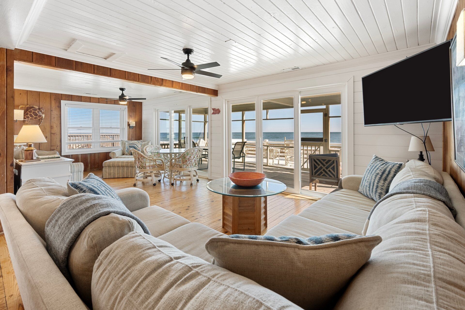 A spacious living room with a large sofa, a coffee table, and a television. There is a balcony with a view of the ocean.