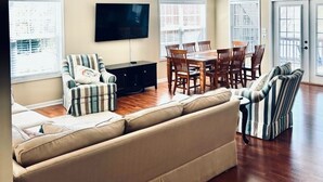 A spacious living room with a large sofa, a dining table with chairs, a flat-screen TV mounted on the wall, and large windows with blinds. - Spacious 5-bedroom lakefront house in Anderson, 13 miles to Clemson (Anderson)