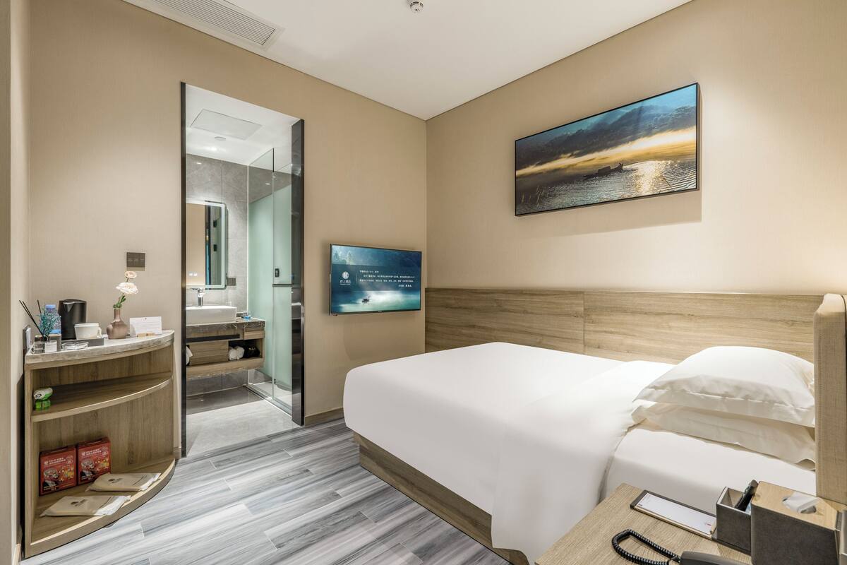 A hotel room with a bed, a desk with a phone, a mirror, a TV, and a painting on the wall.