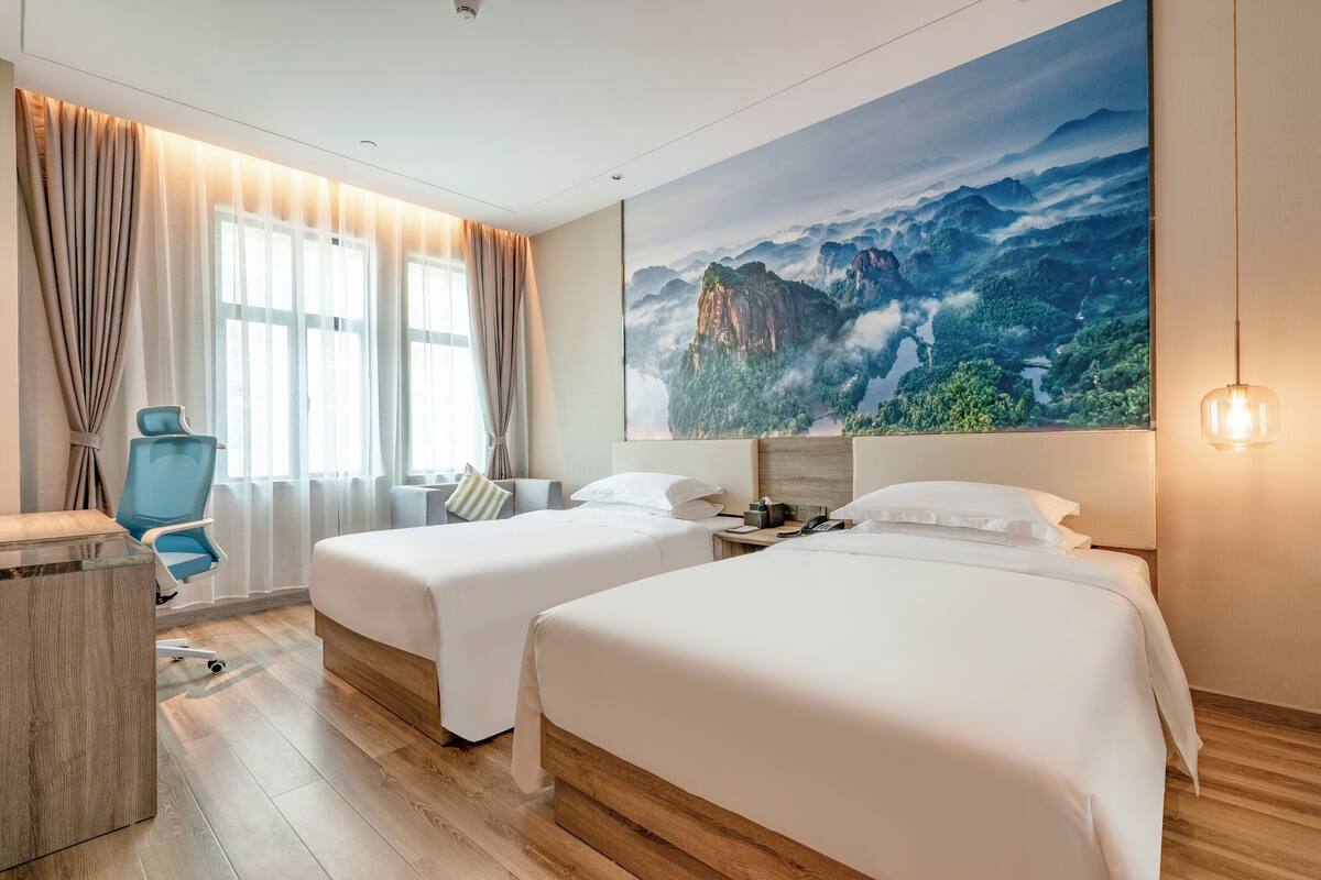A hotel room with two beds, a desk, a chair, and a large mountain view painting.