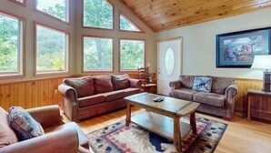 A cozy living room with wooden ceiling, large windows, and a view of greenery. - Expansive home with arcade game, pool table, & Great Smoky Mountains view (Gatlinburg)