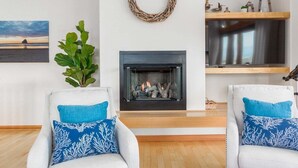 Ein Wohnzimmer mit Kamin, zwei weiße Sessel mit blauen Kissen, einem an der Wand befestigten Fernseher und einem Bilderrahmen mit einer Strandansicht.
