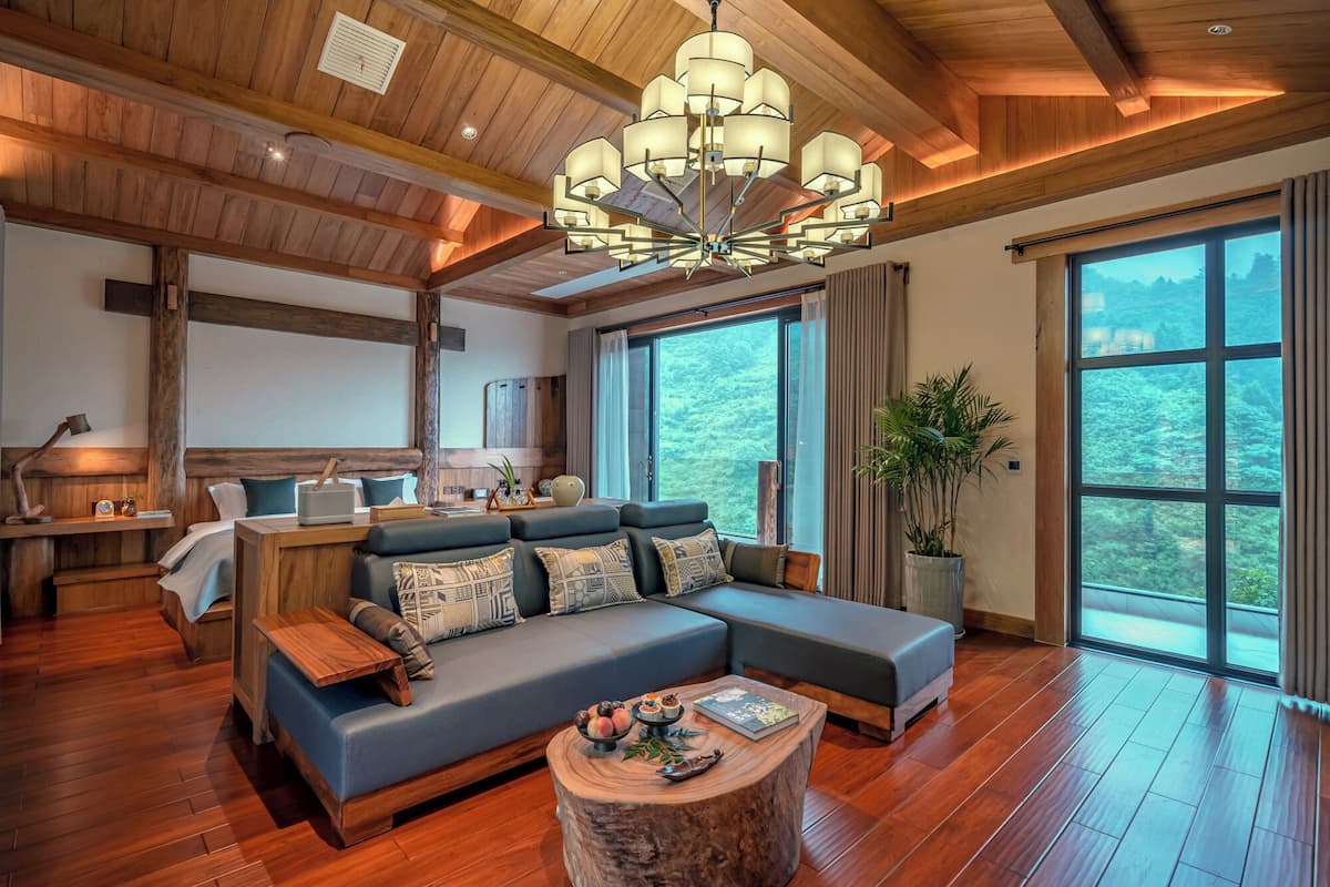 A spacious living room with a large sectional sofa, wooden flooring, a wooden ceiling with exposed beams, and a large window offering a view of the outdoors.