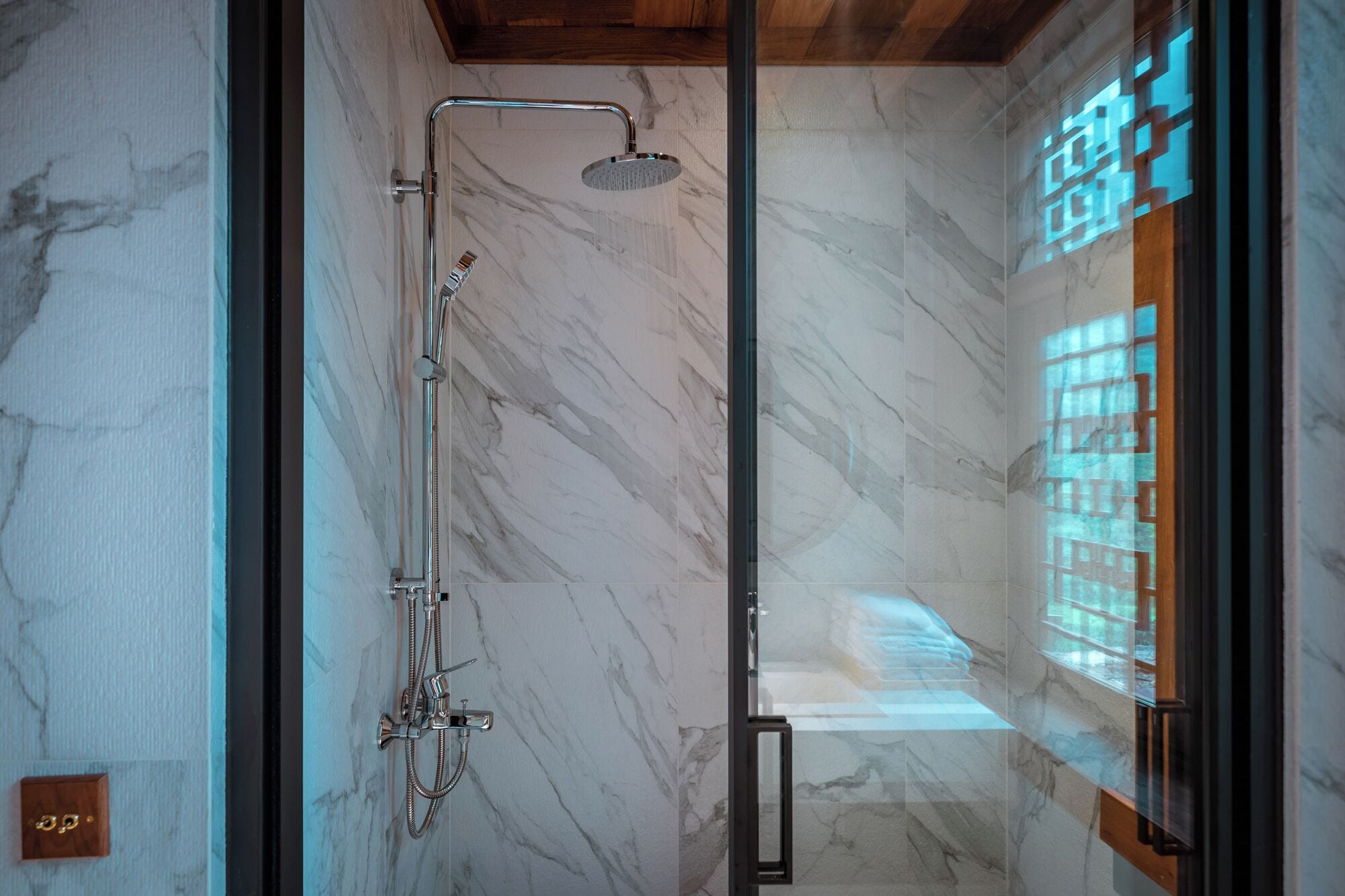 A modern bathroom with a glass shower enclosure, marble walls, and a visible wooden ceiling.