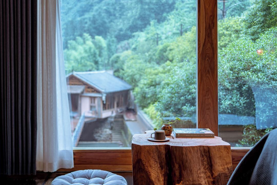 A room with a large window overlooking a traditional building and lush greenery. A wooden table with a book and a cup is visible.