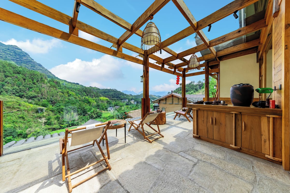 A wooden balcony with a view of a mountainous landscape, wooden furniture, and a large vase on the counter.