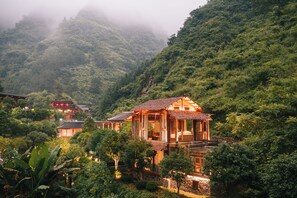 A traditional wooden building with a thatched roof, surrounded by lush greenery and a mountainous backdrop. - Home away from home (Zhangjiajie)