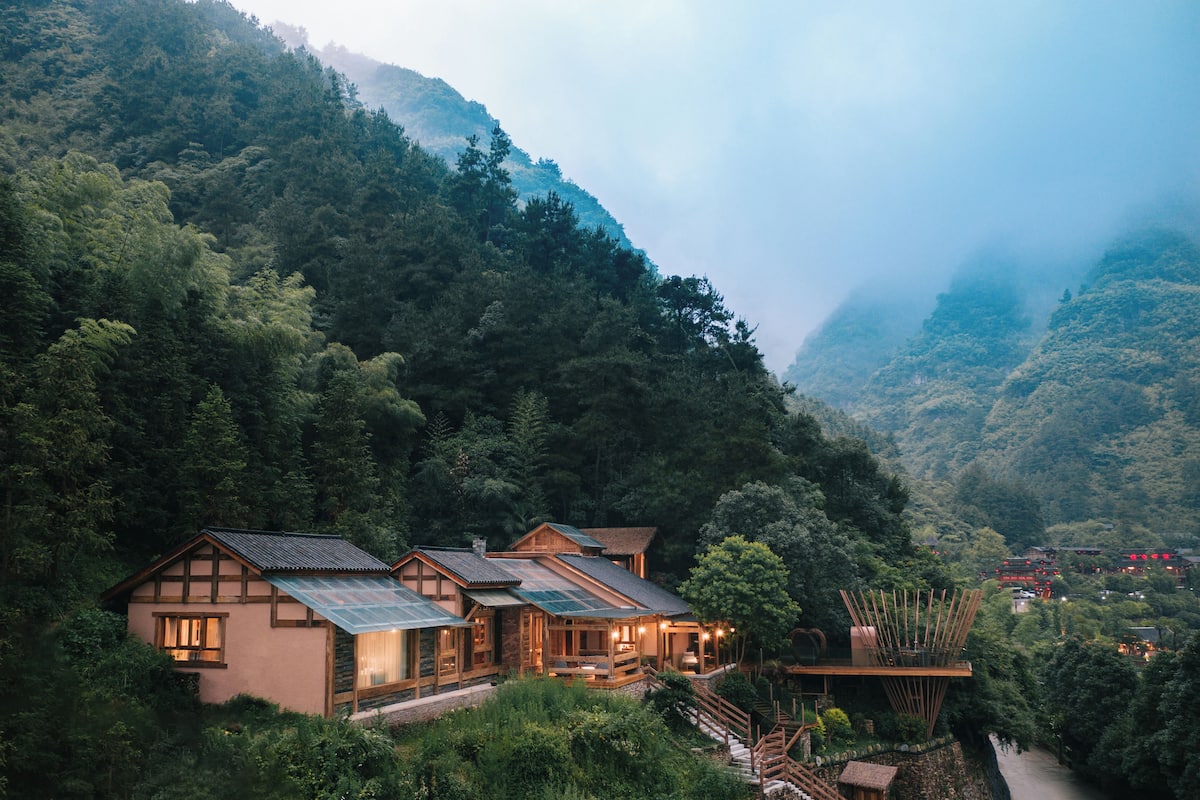 A lodge with multiple wooden cabins nestled in a forested valley, surrounded by steep hills and a river.