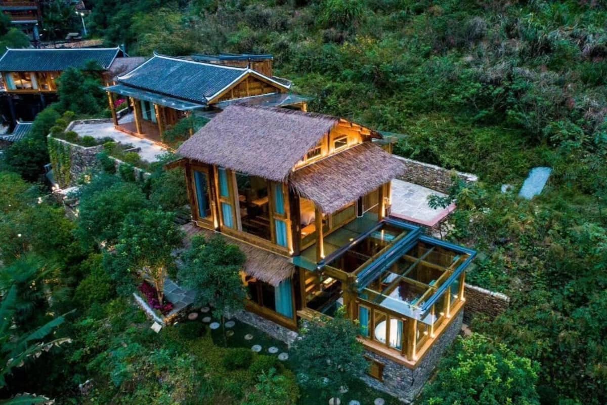 A multi-story wooden house with a thatched roof, surrounded by dense greenery.