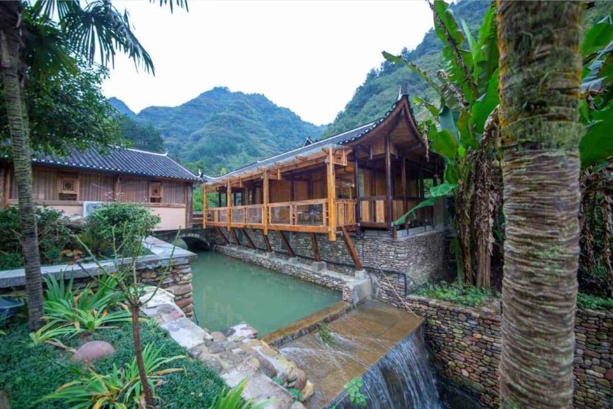 A traditional wooden building with a wooden deck over a water feature, surrounded by lush greenery and mountains in the background.