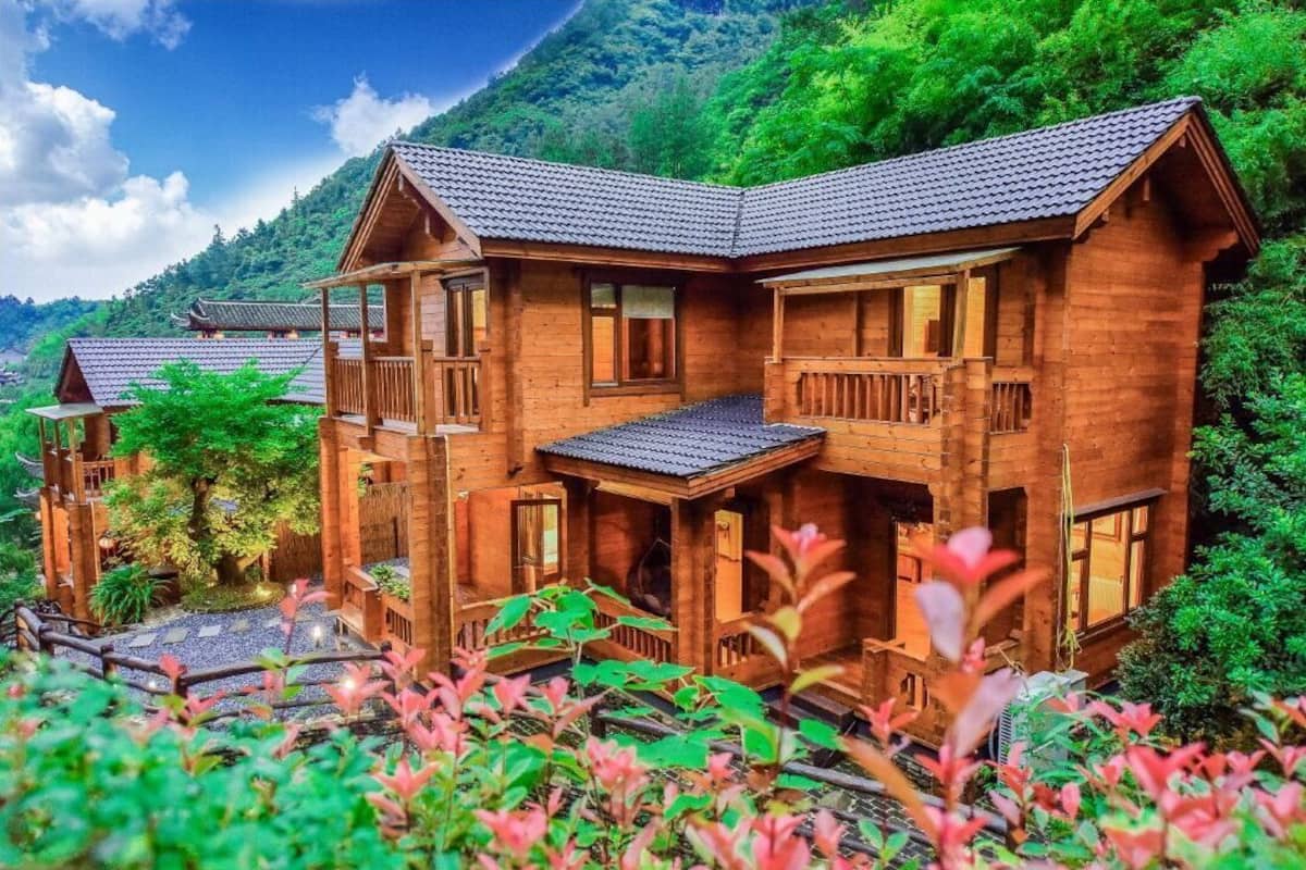 A wooden cabin with a balcony, surrounded by lush greenery and a mountainous backdrop.