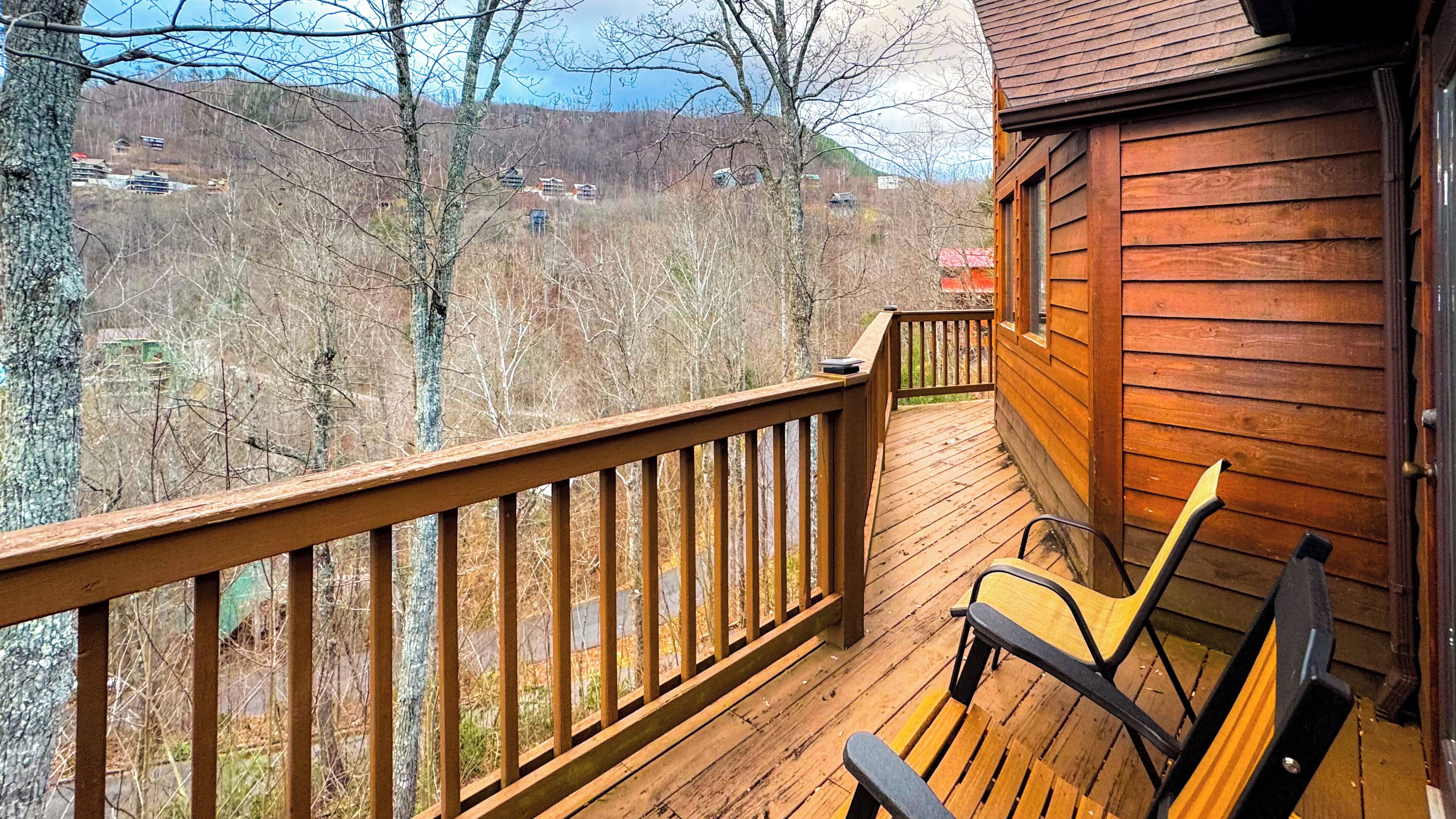 A wooden deck with a bench, overlooking a forested area with buildings in the distance.