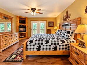 A bedroom with a bed, wooden furniture, a ceiling fan, a fireplace, and a view of the outdoors.