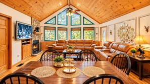 A cozy living room with a wooden ceiling, leather sofas, a dining table with placemats, and a fireplace.