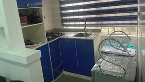 Een kleine keuken met blauwe kastjes, een gasfornuis, een spoelbak en een raam met decoratieve lamellen.