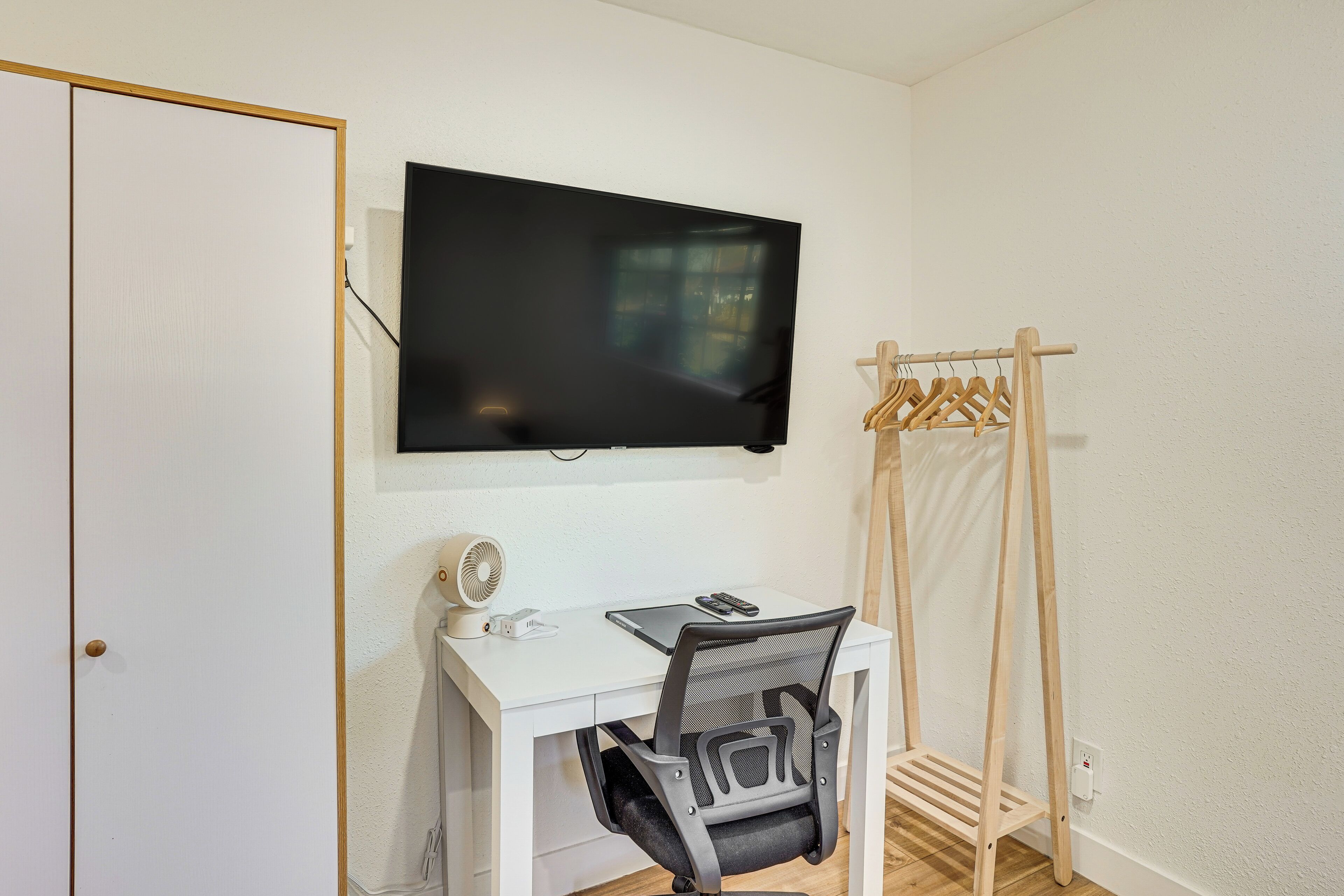A room with a white desk, a mounted TV, a chair, and a wooden drying rack.