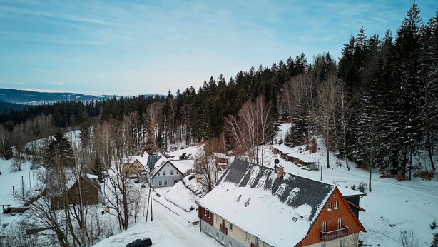 Ein verschneiter Wohnbereich mit Häusern und bewaldeter Landschaft im Hintergrund.
