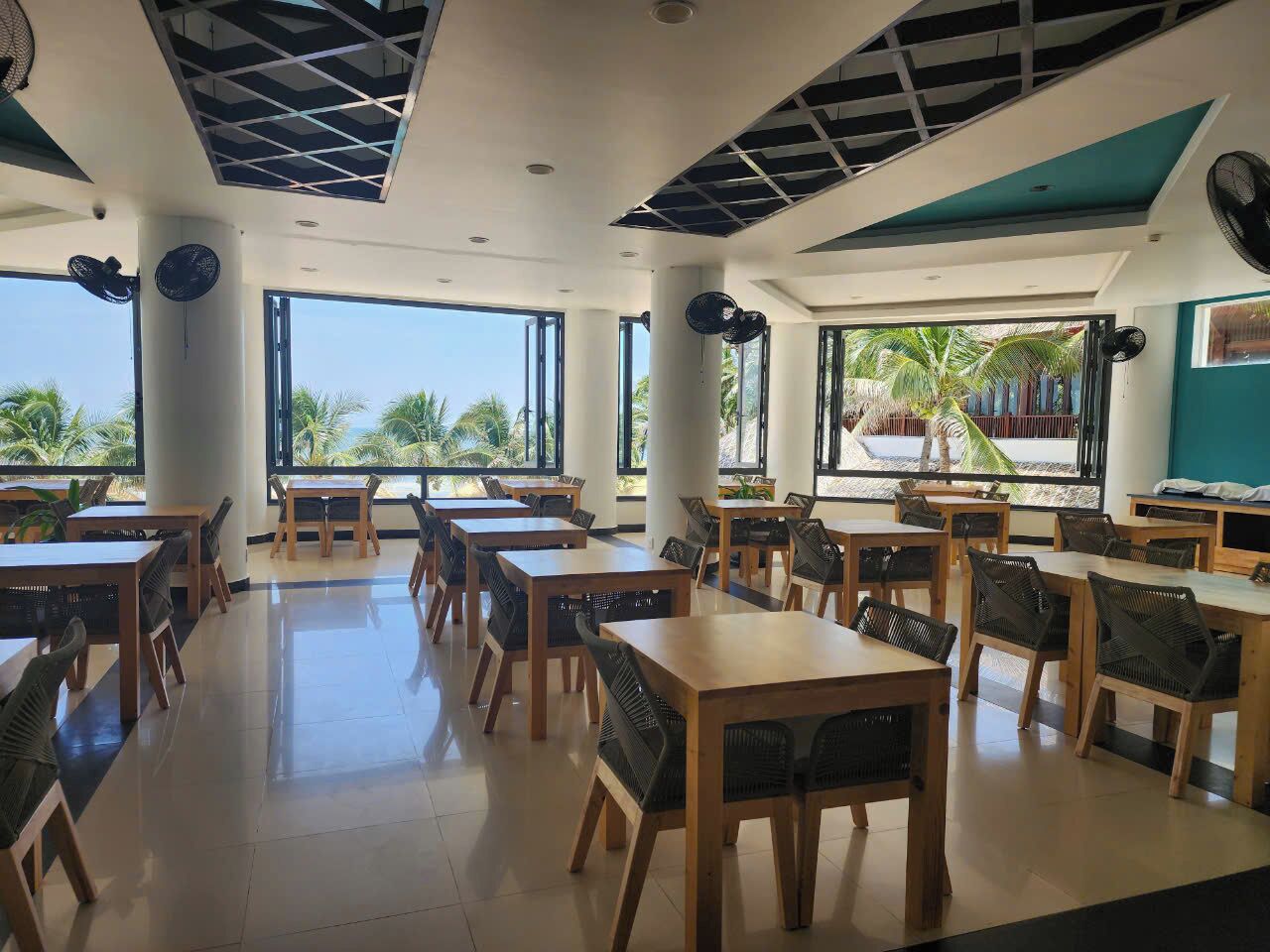 A spacious dining area with wooden tables and chairs, large windows offering views of palm trees, and ceiling fans.