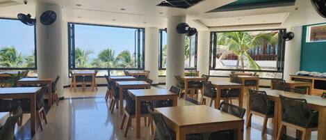 A spacious dining area with wooden tables and chairs, large windows offering views of palm trees, and ceiling fans.