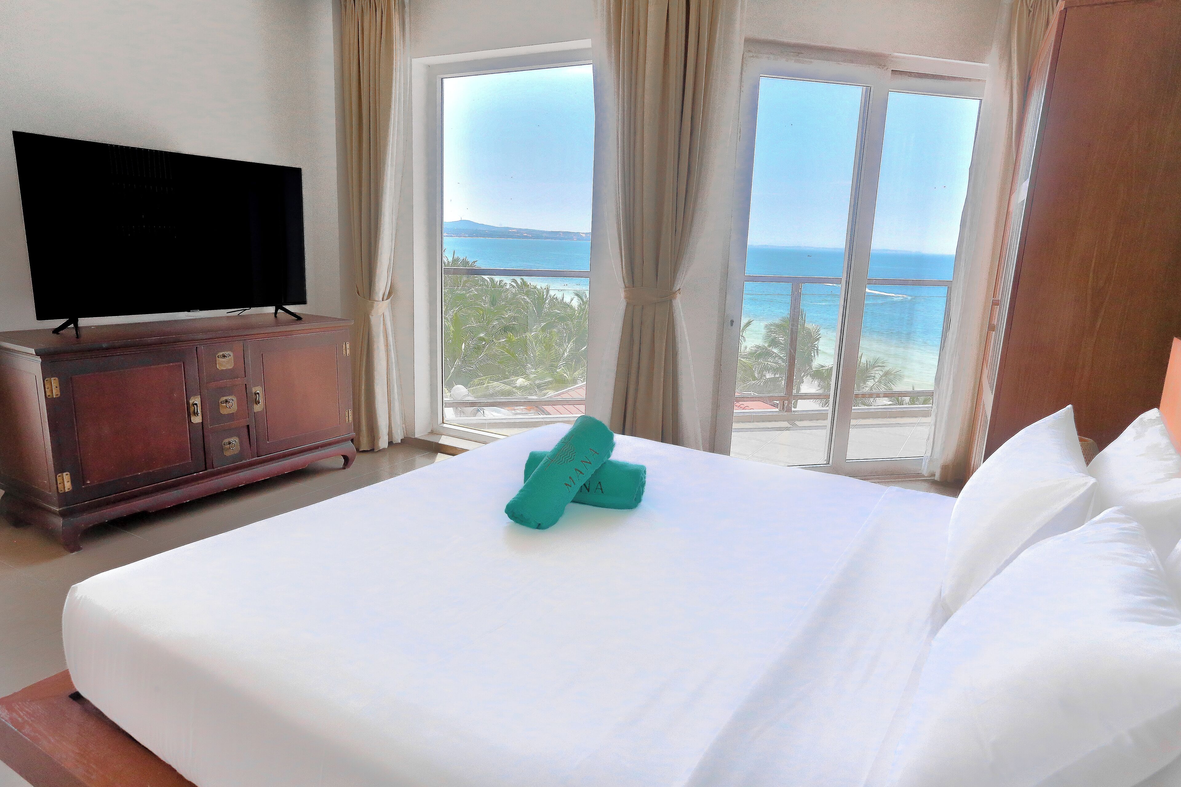 A hotel room with a bed, a television on a wooden cabinet, and a view of the ocean through large windows.