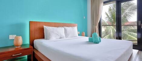 A hotel room with a large bed, a wooden nightstand, a lamp, and a view of palm trees through a window.