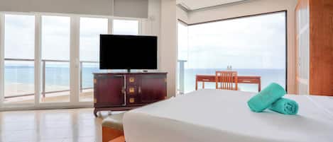 Una habitación de hotel con una cama grande, un televisor de pantalla plana, un escritorio de madera y vista al océano a través de amplias ventanas.