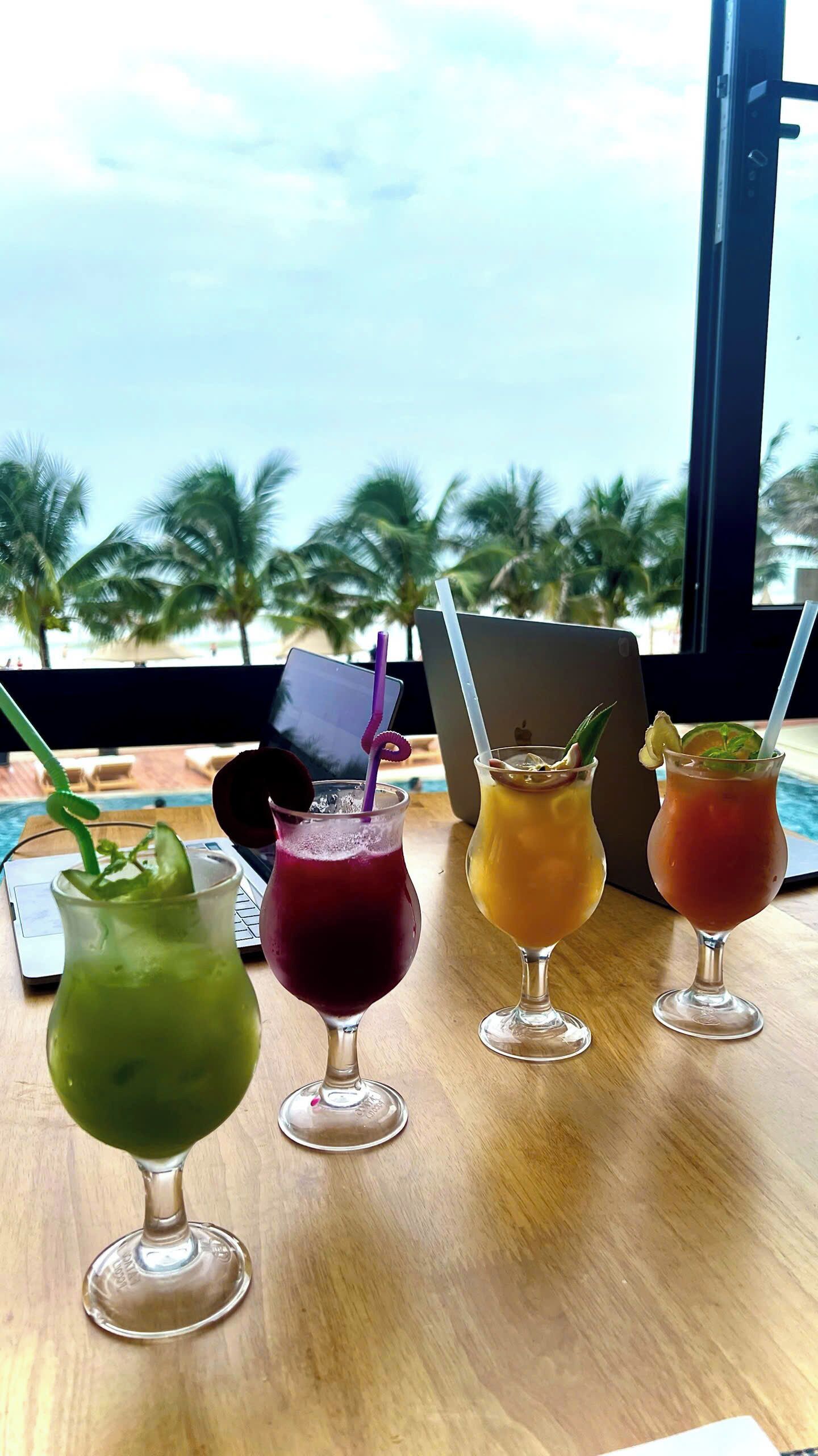 A table with four drinks, a laptop, and a view of palm trees and a pool.
