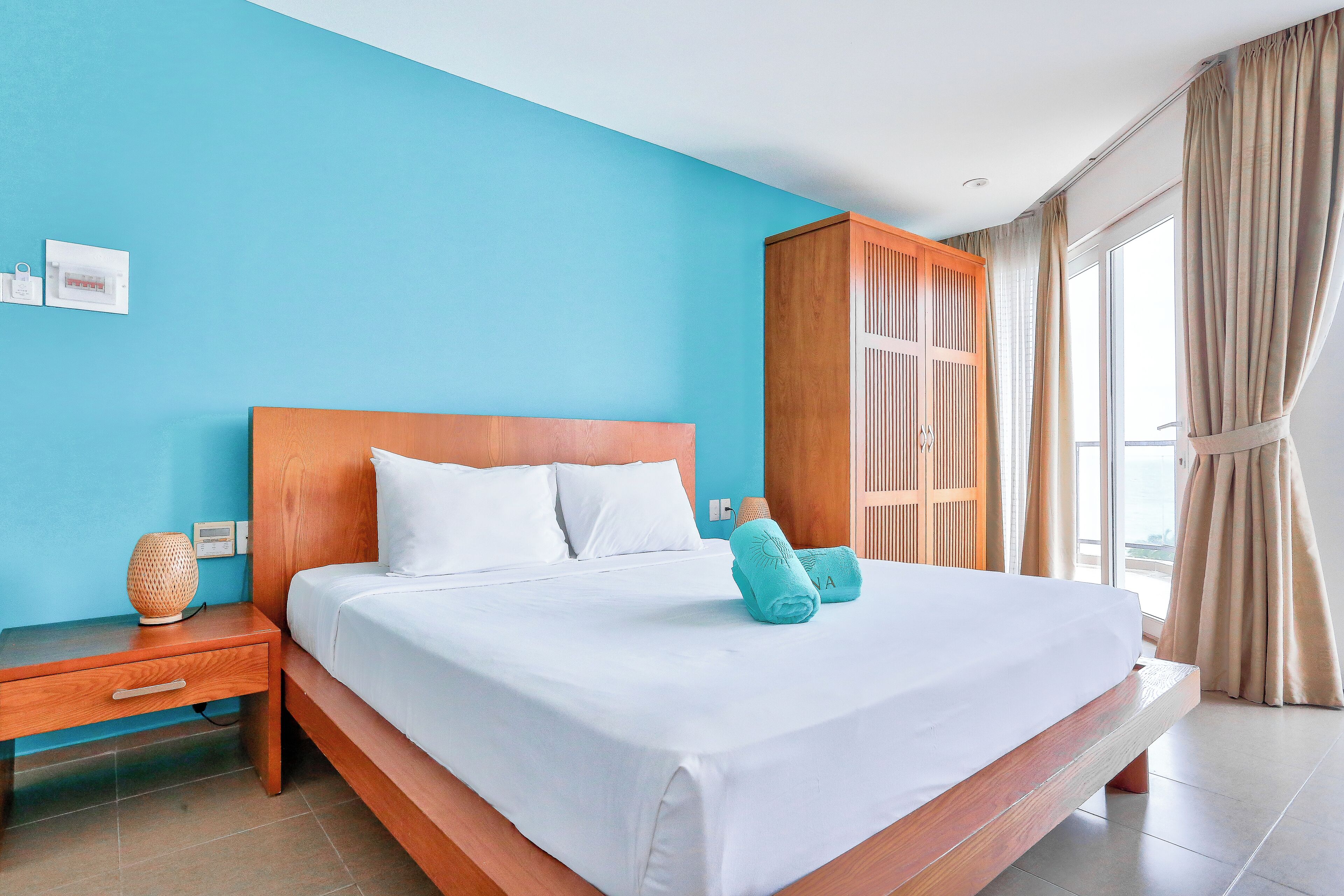 Quarto de hotel com uma cama grande, uma mesa de cabeceira de madeira, um guarda-roupa de madeira e uma janela com cortinas.