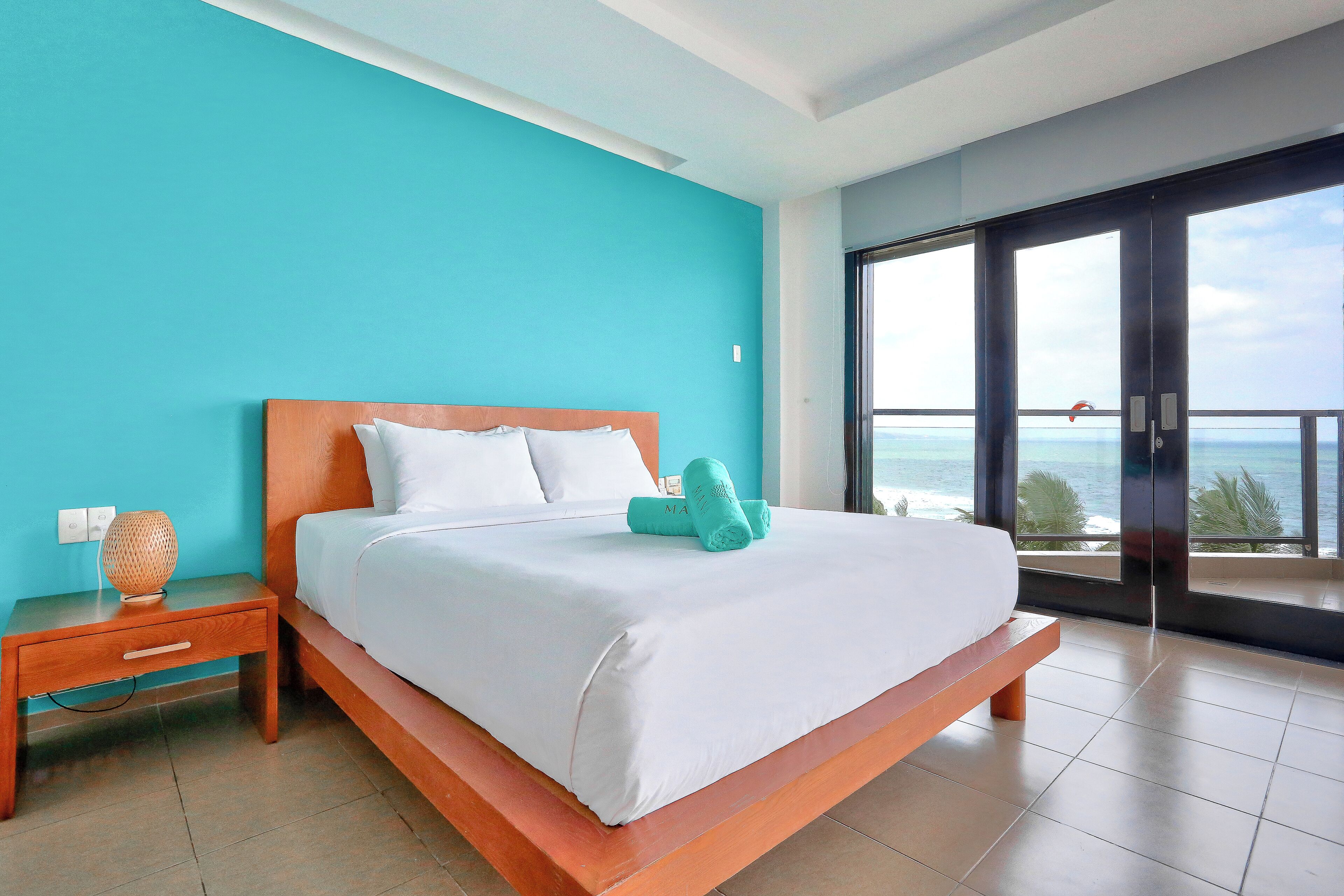 A bedroom with a bed, a nightstand, a window with a view of the ocean, and a balcony.