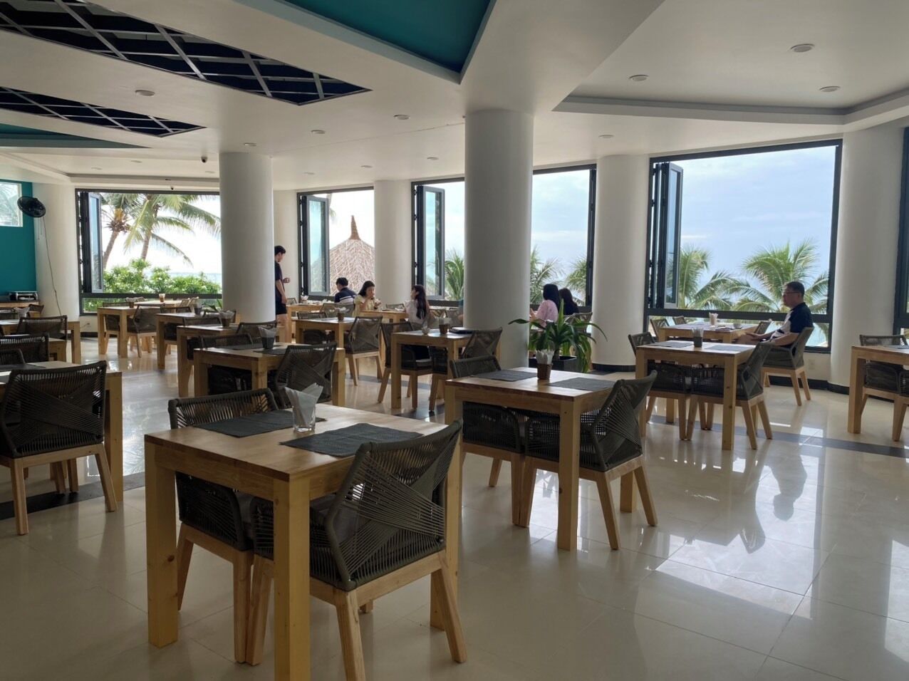 A spacious dining area with wooden tables and chairs, large windows offering views of palm trees and the ocean.