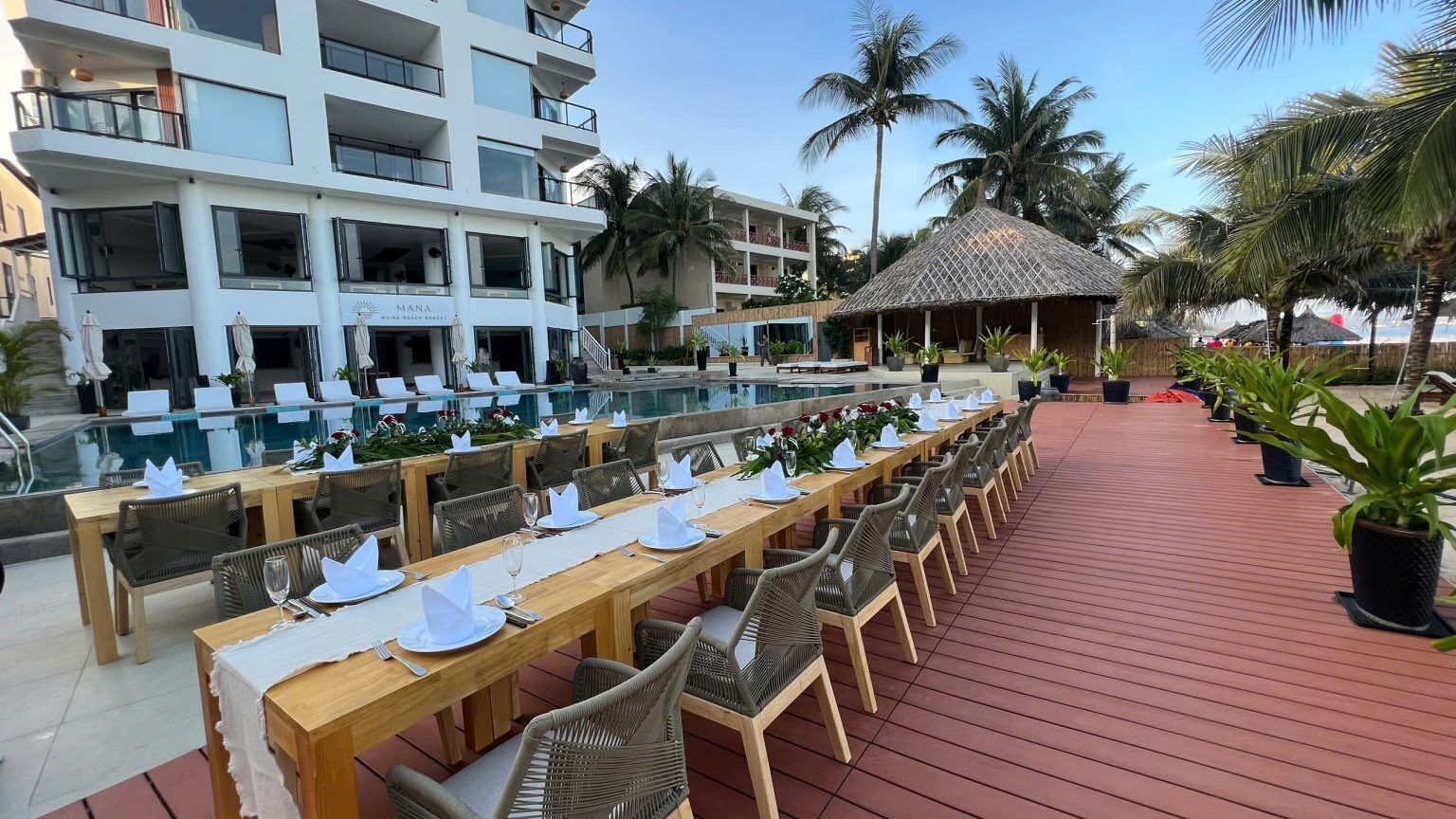 A dining area with long wooden tables and wicker chairs, a pool, and a building with balconies.