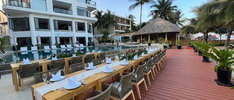 A dining area with long wooden tables and wicker chairs, a pool, and a building with balconies.