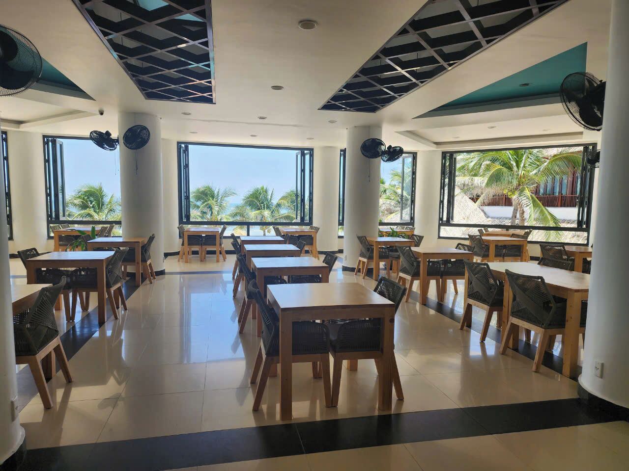 A spacious indoor dining area with multiple tables and chairs, large windows with a view of palm trees, and a ceiling with a geometric pattern.