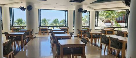 A spacious indoor dining area with multiple tables and chairs, large windows with a view of palm trees, and a ceiling with a geometric pattern.
