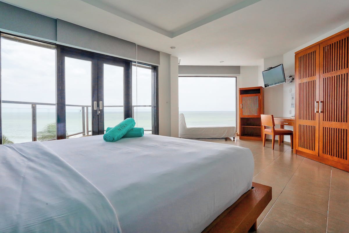 Ein Hotelzimmer mit einem großen Bett, einem Schreibtisch und einem Fernseher. Durch die Fenster ist der Blick auf das Meer.