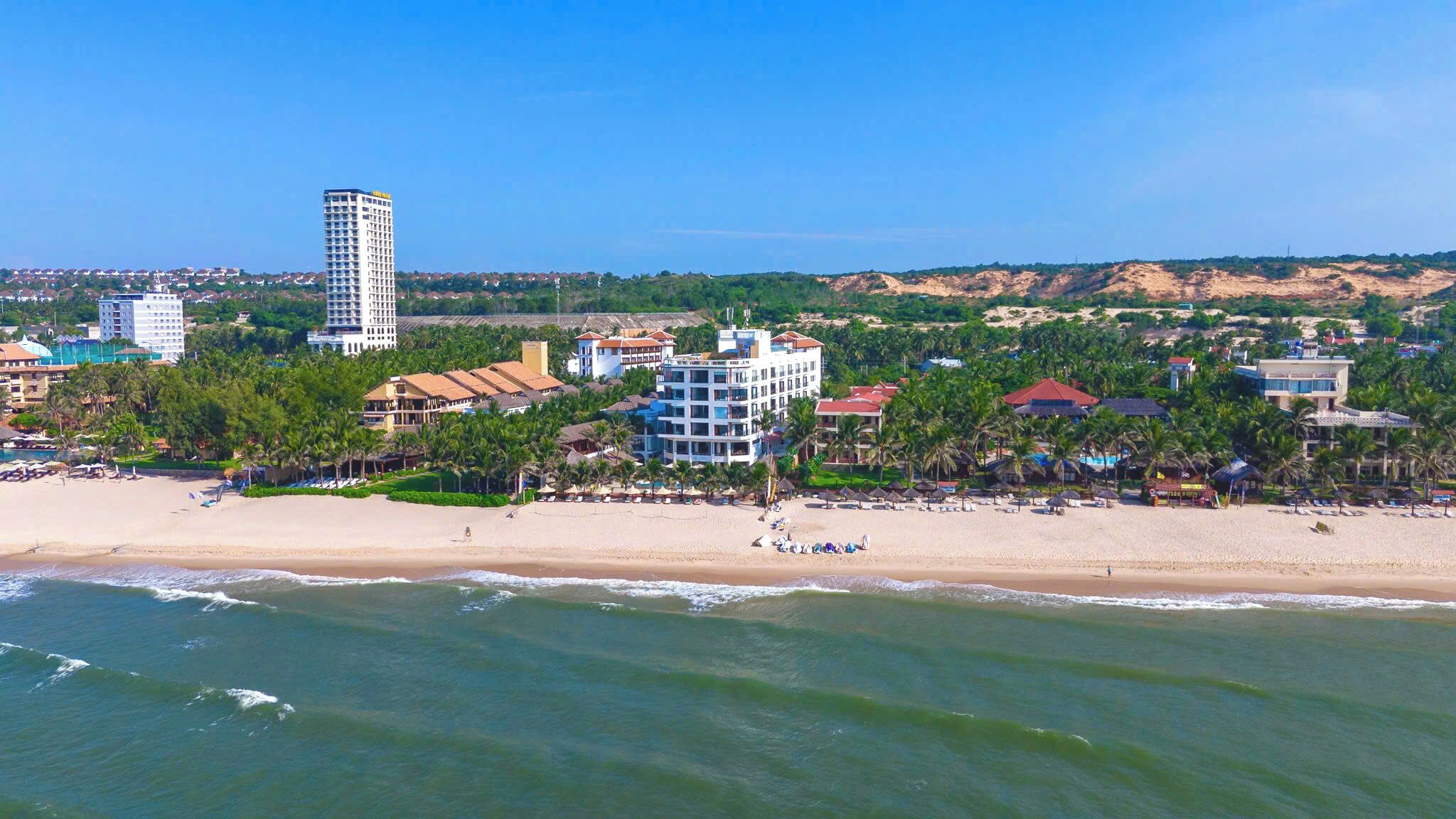 Un complejo turístico frente al mar con villas en la playa, un hotel alto y palmeras.