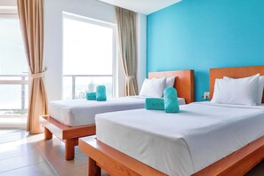 Habitación de hotel con dos camas, cada una con ropa de cama blanca y almohadas color turquesa, cabeceros de madera y vistas al exterior a través de ventanas grandes.