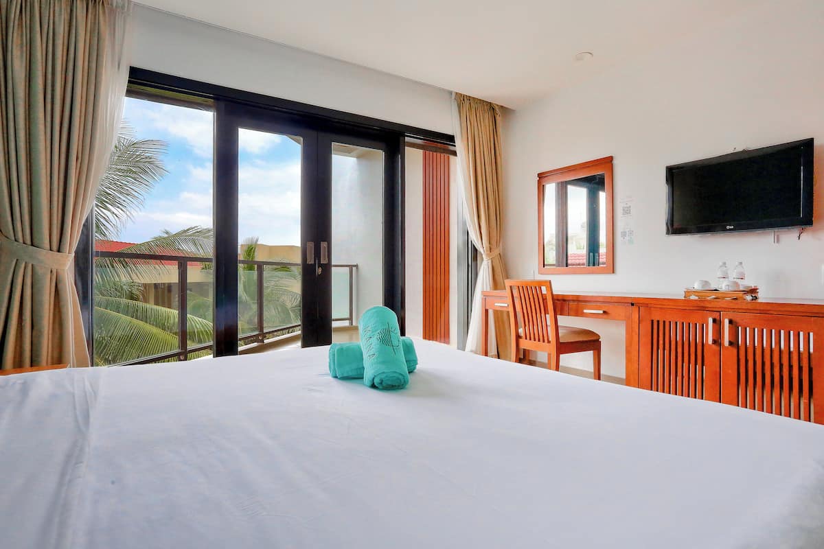 Ein Hotelzimmer mit Bett, Schreibtisch, Stuhl, Fernseher und einem Balkon mit Blick auf Palmen.
