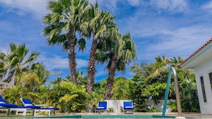 A pool area with palm trees, blue lounge chairs, and a building with a red-tiled roof.