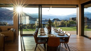 Un comedor con una mesa grande dispuesta para una comida, ventanas amplias con vistas a montañas y un lago, y una decoración de estilo moderno.