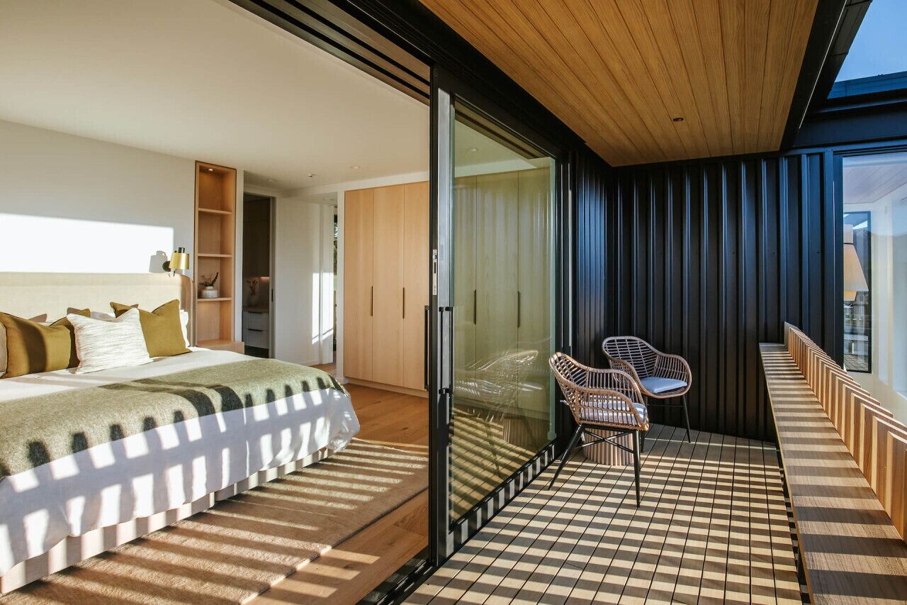 A modern bedroom with a bed, a bedside table, and a chair. There is a sliding glass door leading to a balcony with a checkered floor and a chair.