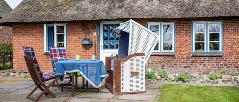 A brick house with a thatched roof, a dining area with a table and chairs, and a small garden.
